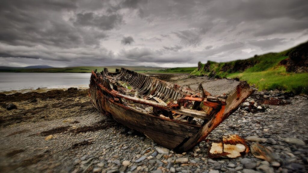 Abandoned wooden shipwreck decaying on a rocky shoreline under dramatic cloudy skies near a calm coastal inlet.