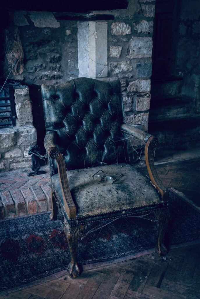 A worn leather armchair sits abandoned beside a cold stone hearth, its surface layered with dust, cobwebs, and age