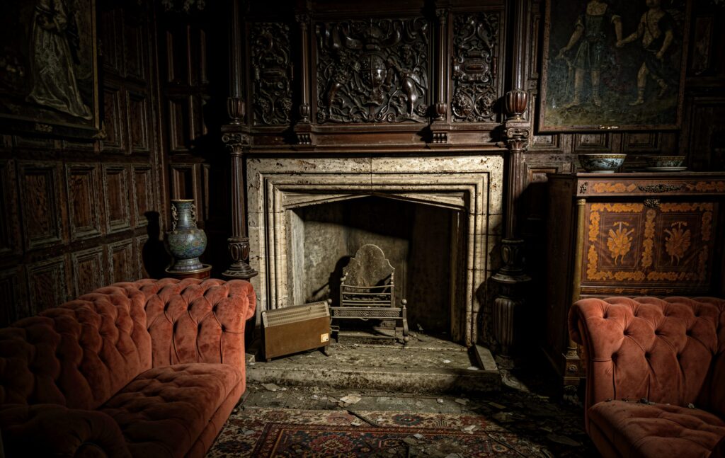 The Cold Hearth. In a dark wood-panelled room, two heavy red Chesterfield sofas face an ornate stone fireplace that no longer holds a fire