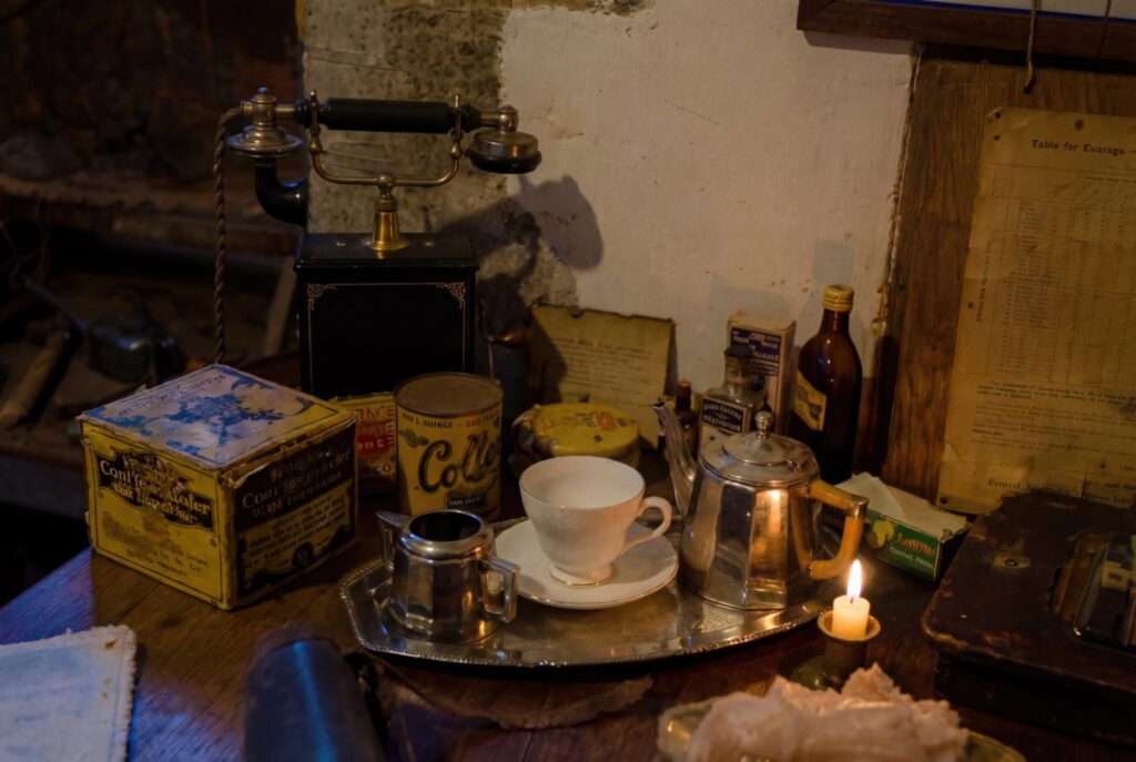 A vintage table setting frozen in time. An antique telephone stands silent beside weathered tins and a silver tea service, all illuminated by the warm, solitary...