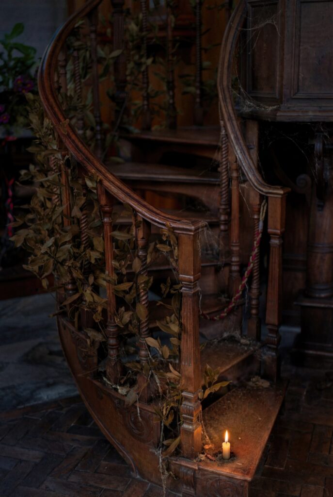 A candle burning at the foot of a decaying wooden staircase, its banister entwined with dead leaves and cobwebs