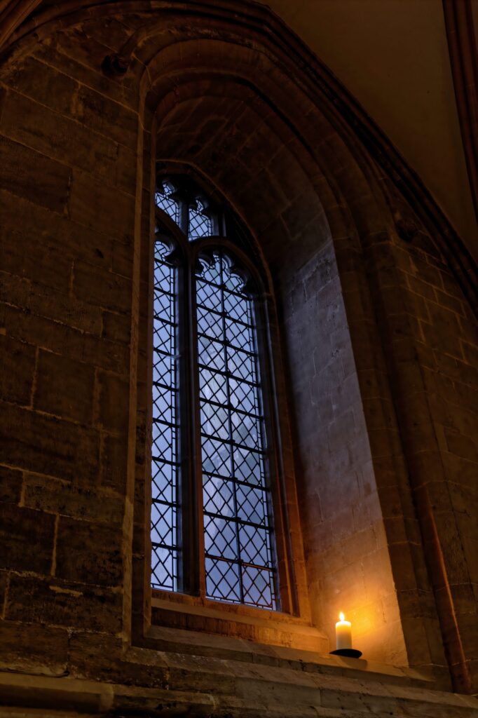 A solitary candle casts a warm, defiant glow against the cold, leaded glass of a Gothic window