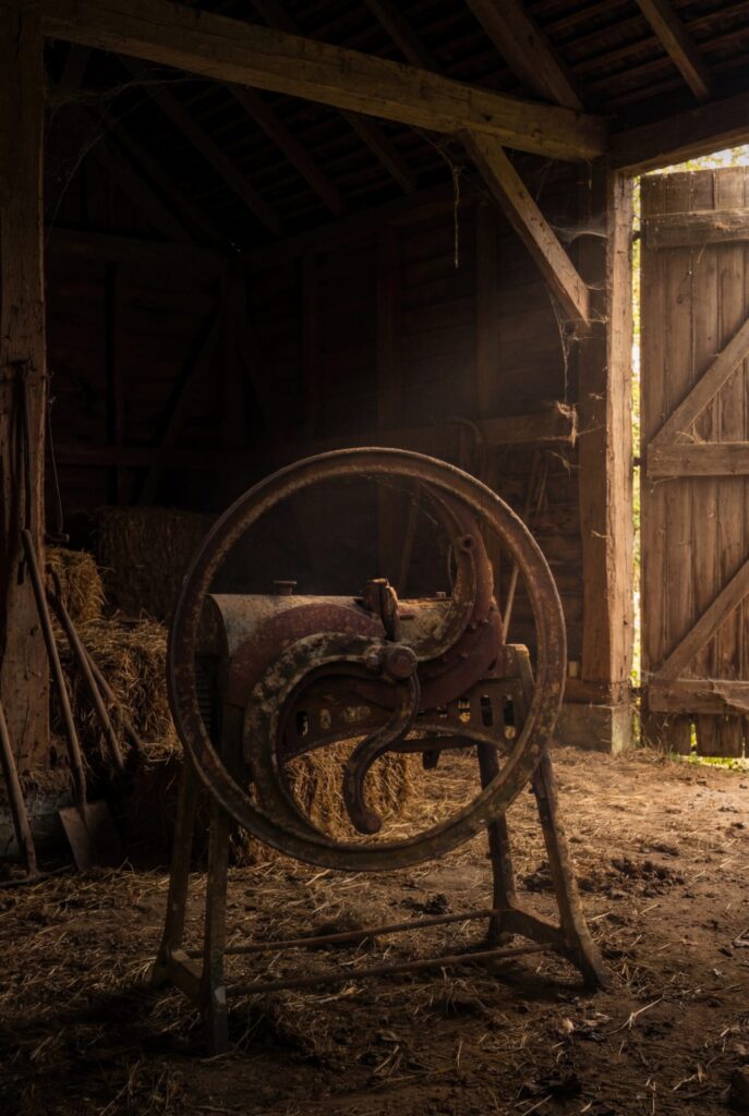 An abandoned hand-operated farm machine stands within a dim wooden barn, surrounded by straw and dust, its rusted wheel bearing the quiet weight of labour, time...