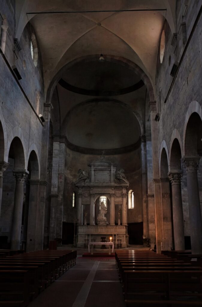 A vast, dimly lit church interior recedes into shadow, its symmetry and stone weight drawing the eye toward a solitary altar light