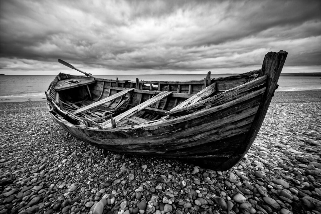 The Wreck on the Shore. A broken wooden boat lies on a pebble beach beneath a heavy sky